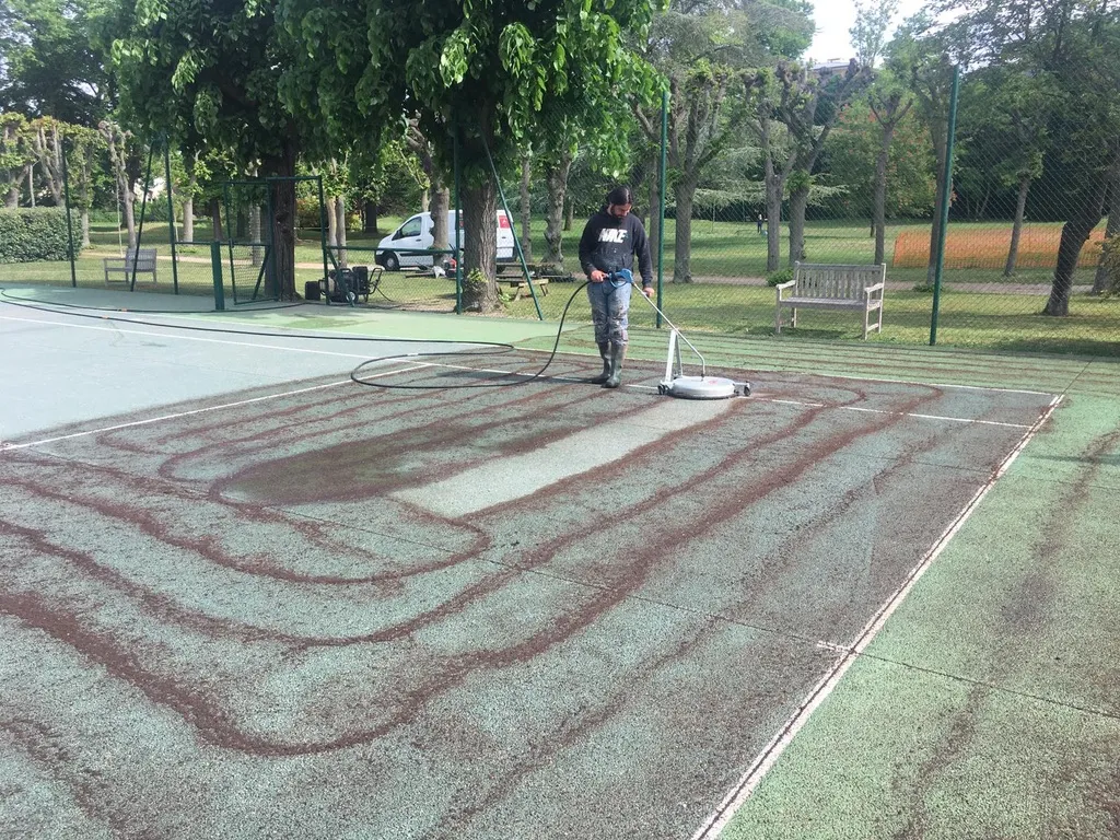 Nettoyage d'un court de tennis avant la rénovation en béton poreux, utilisant un nettoyeur haute pression pour enlever les saletés accumulées.