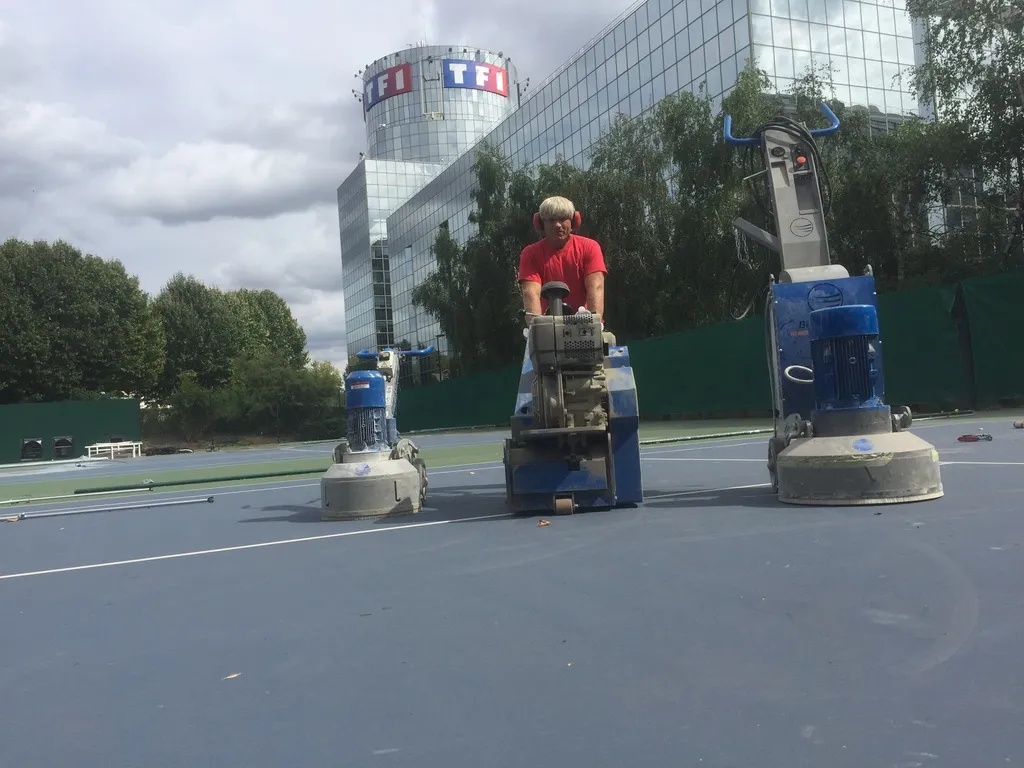 Réparation d'un terrain de tennis avec des machines de ponçage et rabotage, réalisée par un professionnel équipé, en face du siège de TF1.